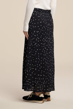 A person is photographed from the back wearing a white long-sleeve top, black sneakers, and the CADENCE SILK SKIRT by Velvet by Graham & Spencer—a black and white polka dot silk maxi skirt.