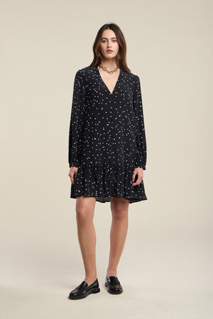 A woman wearing the ELSA SILK DRESS by Velvet by Graham & Spencer—a black, long-sleeve, V-neck dress with white polka dots and a ruffled hem—paired with black loafers and a gold chain necklace.