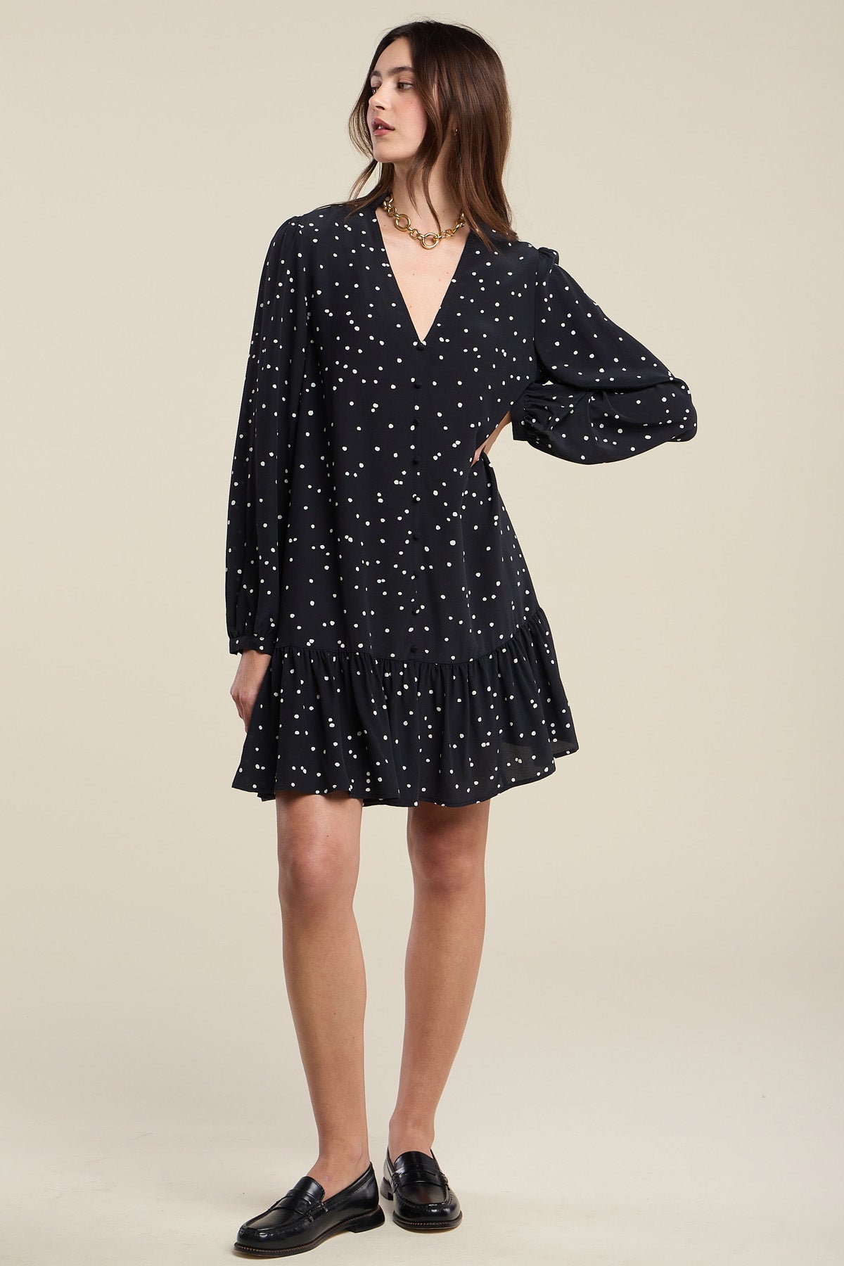 A woman wearing the ELSA SILK DRESS by Velvet by Graham & Spencer—a black, long-sleeve, V-neck dress with white polka dots and a ruffled hem—paired with black loafers, stands against a plain background.-41971008176321