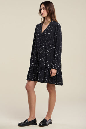 A woman stands in profile wearing the Velvet by Graham & Spencer ELSA SILK DRESS—a loose-fitting, long-sleeve black silk dress with white polka dots and a ruffled hem—paired with black loafers.