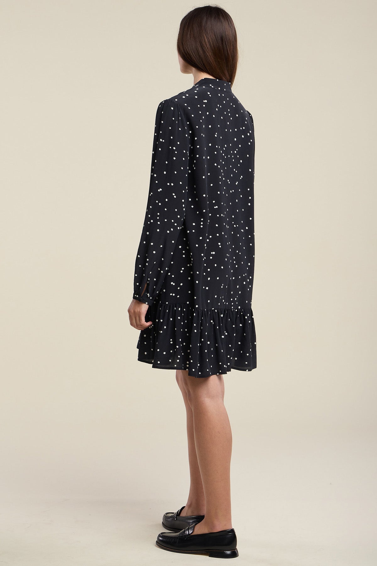 A woman stands facing away against a plain beige background, wearing the ELSA SILK DRESS by Velvet by Graham & Spencer—a black long-sleeve dress with white polka dots and a ruffle hem—paired with black loafers.