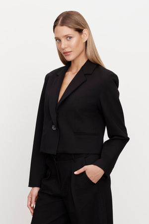 A person with long, straight hair wearing the ANYA PONTE CROPPED BLAZER from Velvet by Graham & Spencer and black pants stands against a plain background with one hand in their pocket.