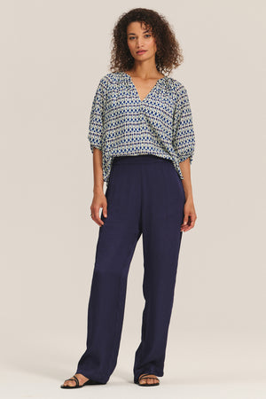 A woman wears the HARMONY BLOUSE by Velvet by Graham & Spencer—a flowy, printed cotton voile top with elbow-length sleeves—paired with loose navy trousers and black sandals, standing against a plain light background.