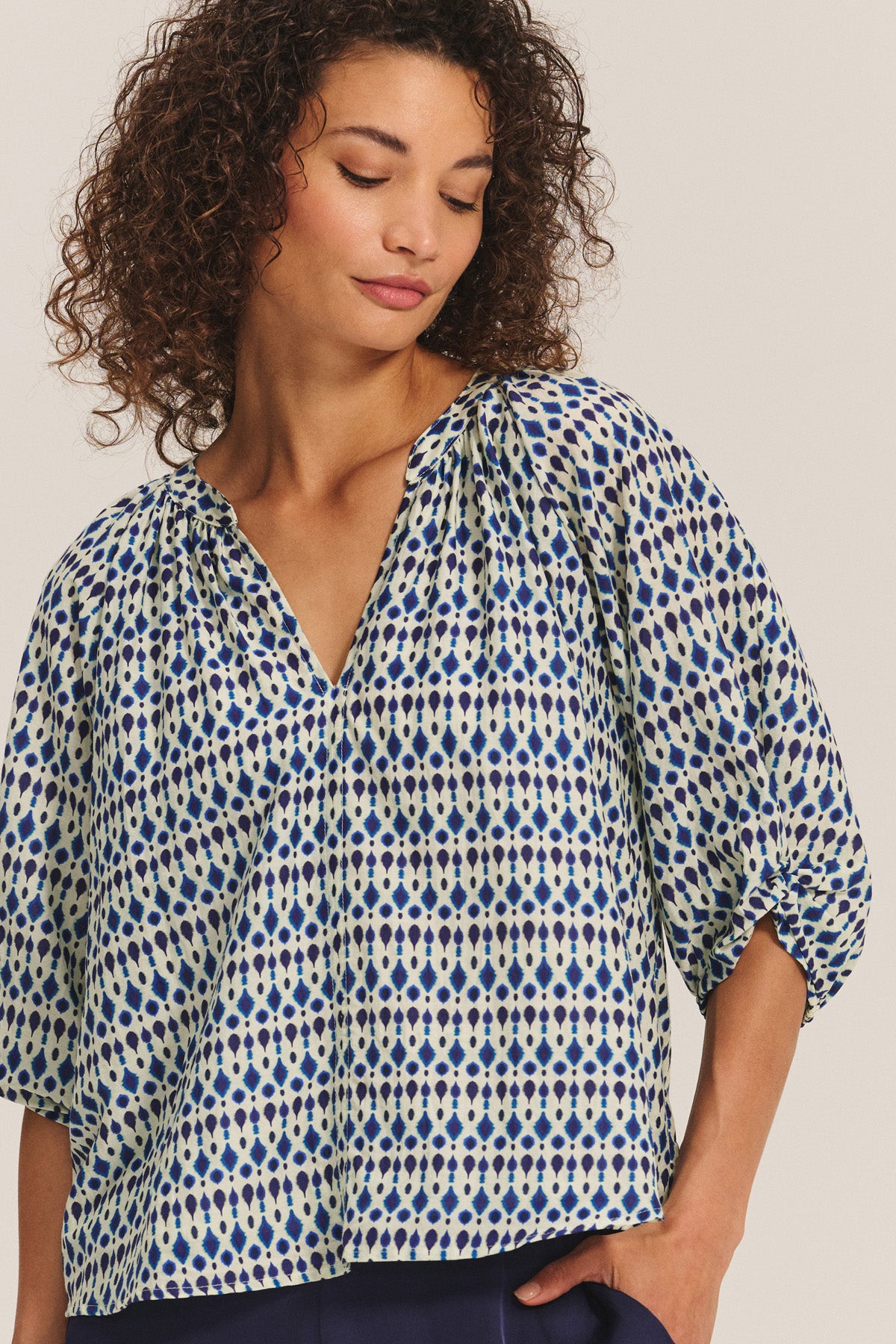A woman wears the HARMONY BLOUSE by Velvet by Graham & Spencer—a flowy printed cotton voile top with blue and beige geometric shapes and three-quarter sleeves—against a neutral background.-40732814868673