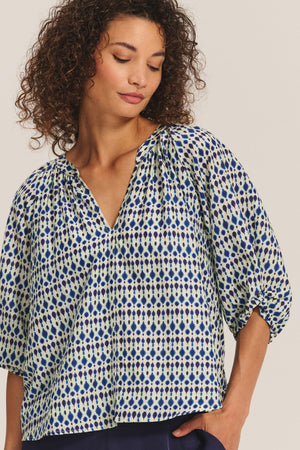 A woman wears the HARMONY BLOUSE by Velvet by Graham & Spencer—a flowy printed cotton voile top with blue and beige geometric shapes and three-quarter sleeves—against a neutral background.