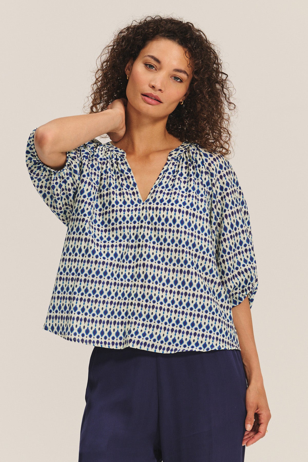 A woman stands with one hand behind her neck, wearing the HARMONY BLOUSE by Velvet by Graham & Spencer—a flowy, patterned cotton voile top with blue and cream dots—paired with navy wide-leg pants.-40732814901441