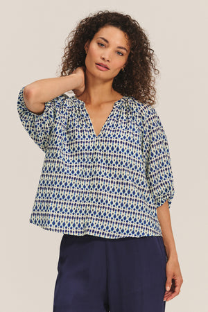 A woman stands with one hand behind her neck, wearing the HARMONY BLOUSE by Velvet by Graham & Spencer—a flowy, patterned cotton voile top with blue and cream dots—paired with navy wide-leg pants.