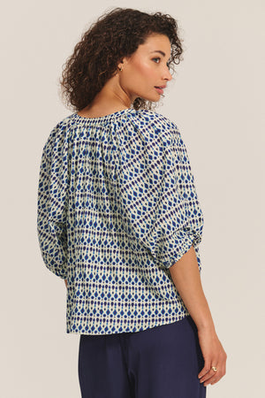 A woman with curly hair wears Velvet by Graham & Spencer's HARMONY BLOUSE, a blue and white printed cotton voile top, paired with dark pants against a light background, looking over her shoulder.