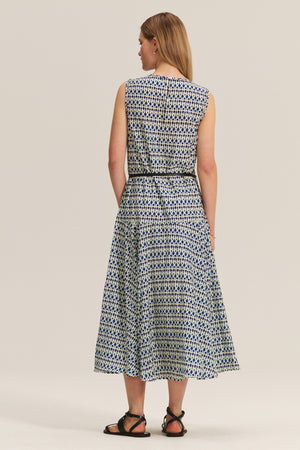 A woman faces away, wearing the KAITLYN DRESS by Velvet by Graham & Spencer—a sleeveless silk cotton voile midi dress with a detachable belt—paired with black sandals, set against a plain background.