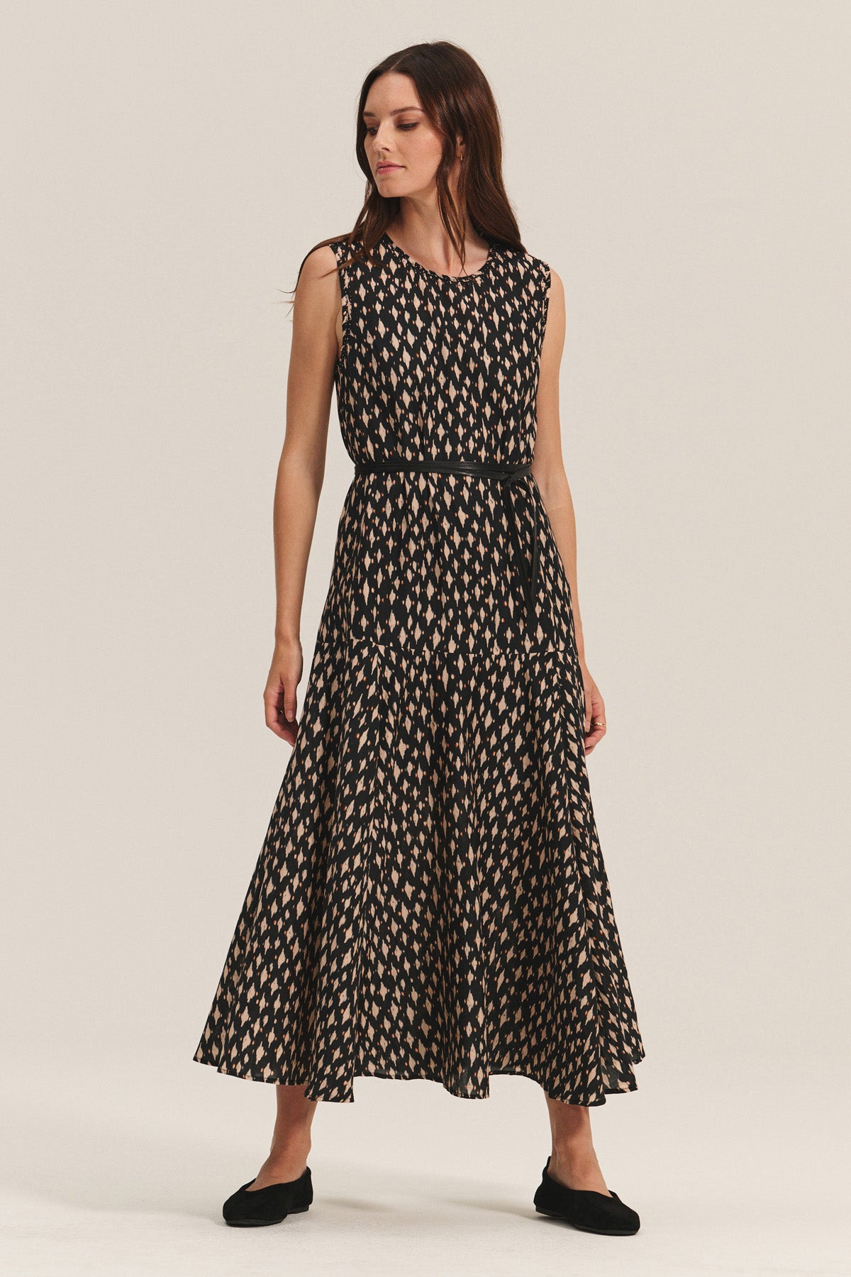 A woman stands against a plain background wearing the KAITLYN DRESS by Velvet by Graham & Spencer, a sleeveless, ankle-length black printed silk cotton voile dress with a beige geometric pattern, detachable waist belt, and black flats.-40248070340801