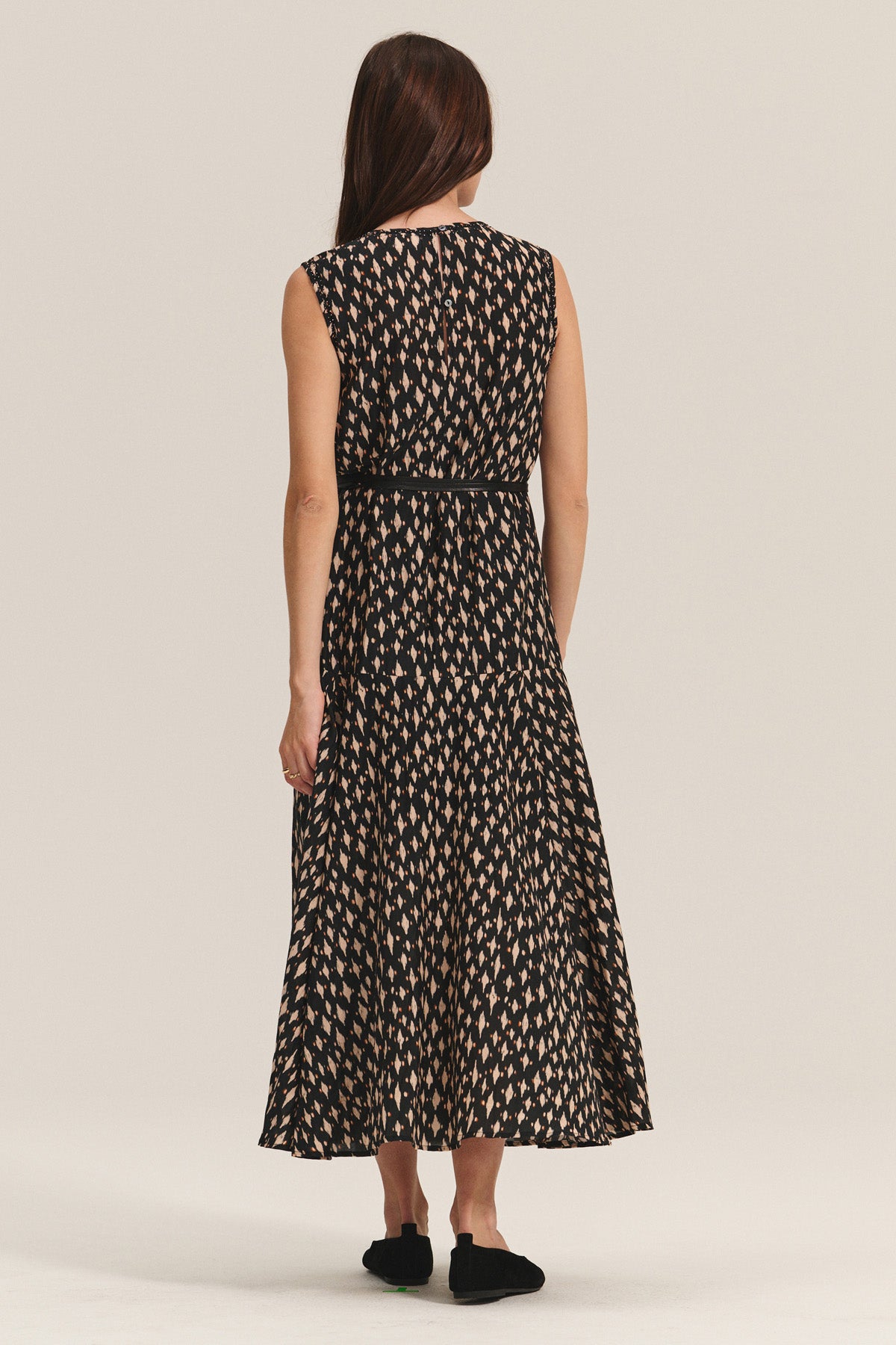 A woman faces away, wearing the KAITLYN DRESS by Velvet by Graham & Spencer—a sleeveless, ankle-length black printed silk cotton voile dress with a beige geometric pattern—paired with black shoes.