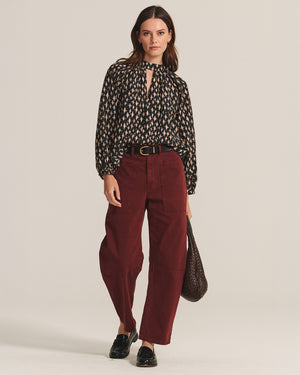 A woman in the MALIA BLOUSE by Velvet by Graham & Spencer, burgundy wide-leg pants, black loafers, and holding a dark woven bag stands against a plain background.