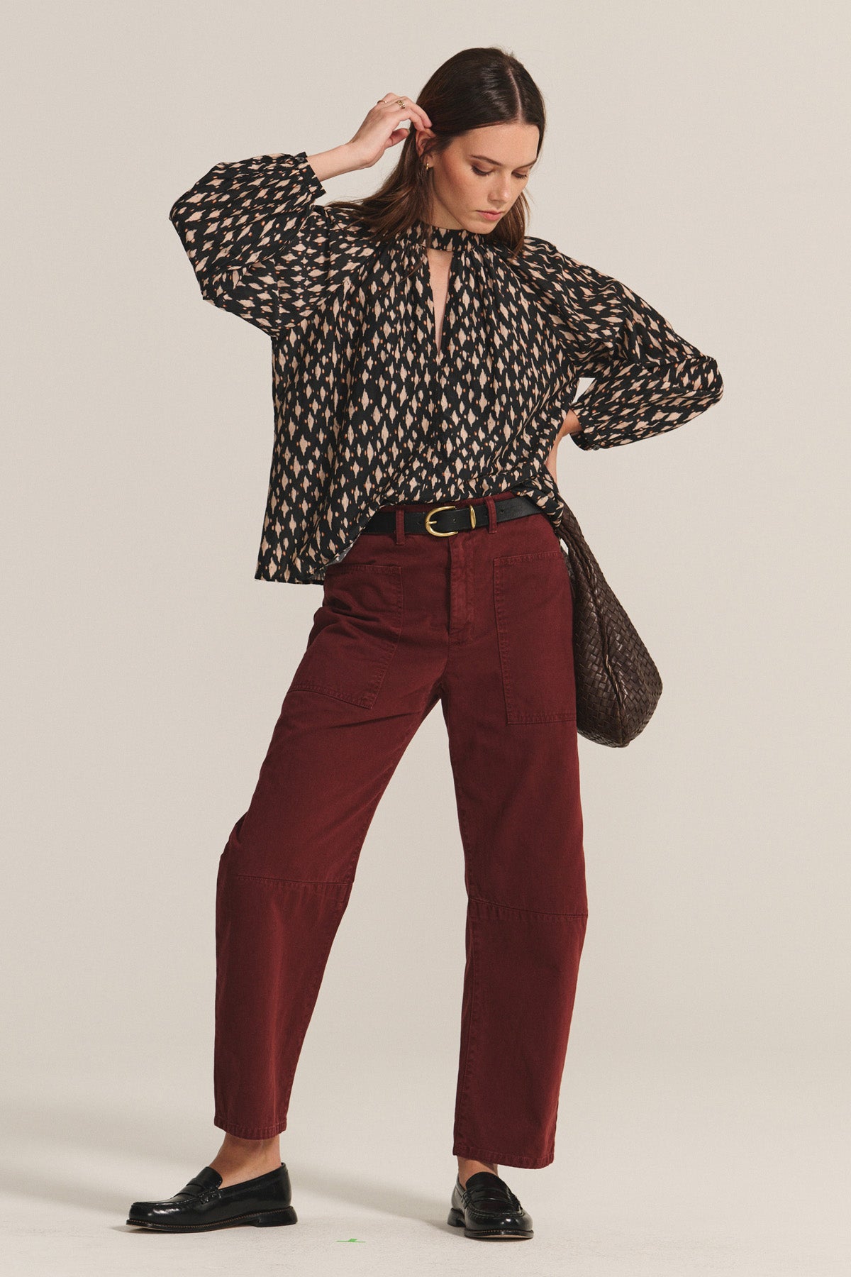 A woman wears a patterned long-sleeve blouse, Velvet by Graham & Spencer BRYLIE SANDED TWILL UTILITY PANT in maroon, black loafers, black belt, and carries a brown shoulder bag.