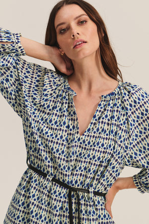 A woman poses against a neutral background wearing the RAINE DRESS by Velvet by Graham & Spencer, a patterned silk cotton voile midi with a V-neckline, subtle pleats, and a thin black belt.