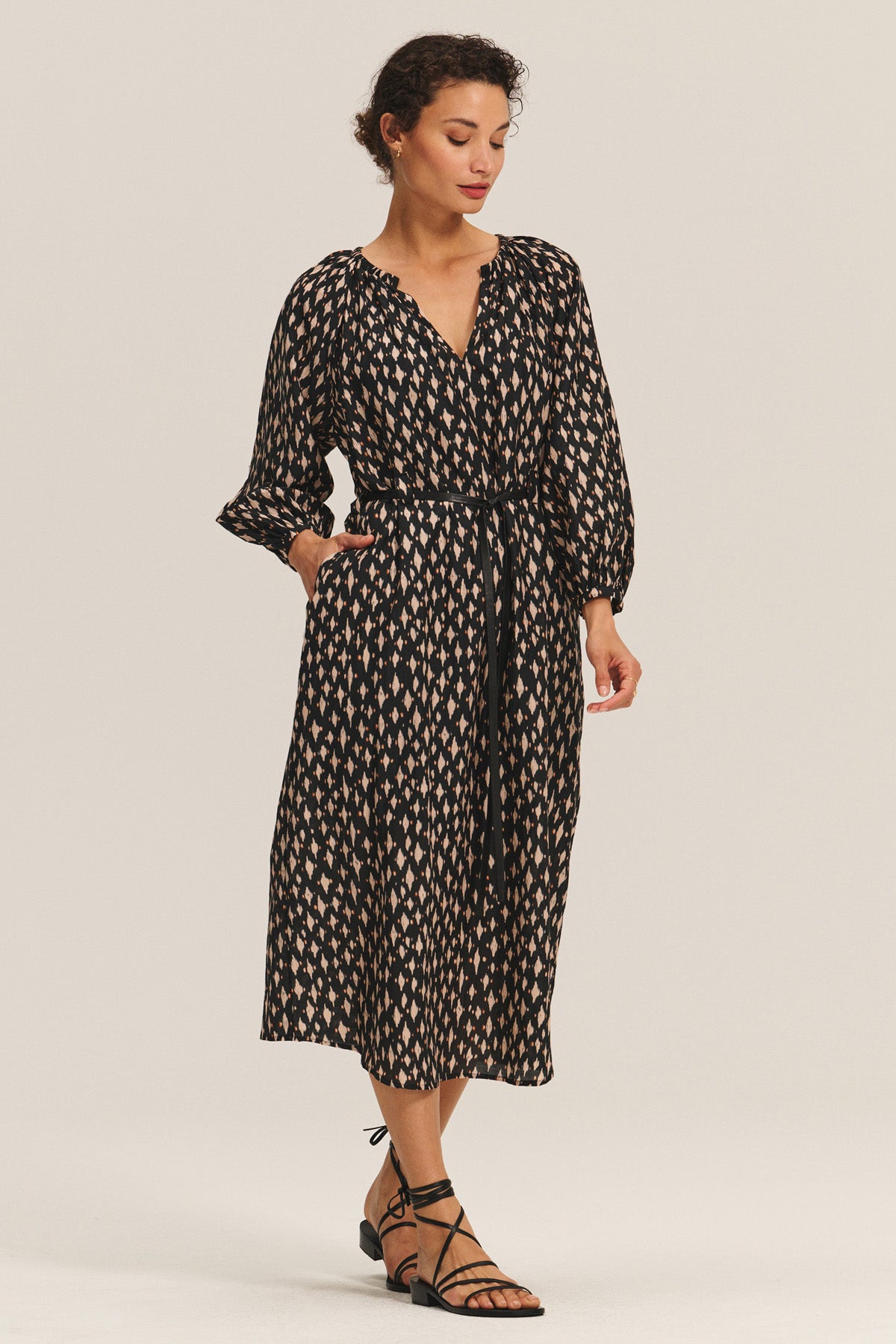 A woman wears the RAINE DRESS by Velvet by Graham & Spencer—a long-sleeve black silk cotton voile midi with pleats, waist tie, and pockets—paired with black strappy sandals.-40688718315713