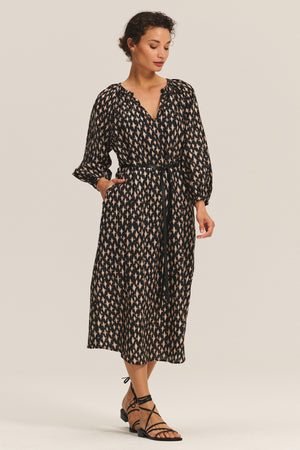 A woman wears the RAINE DRESS by Velvet by Graham & Spencer—a long-sleeve black silk cotton voile midi with pleats, waist tie, and pockets—paired with black strappy sandals.