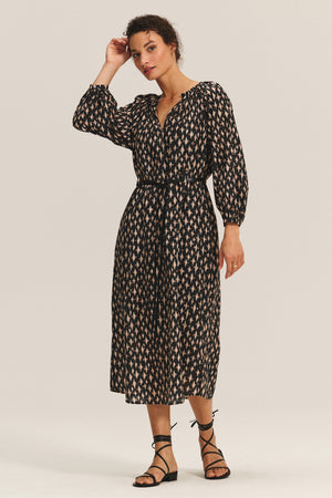 A woman wears the RAINE DRESS by Velvet by Graham & Spencer—a long-sleeve, patterned silk cotton voile midi dress with pleats and a belted waist—styled with black strappy sandals against a plain light background.