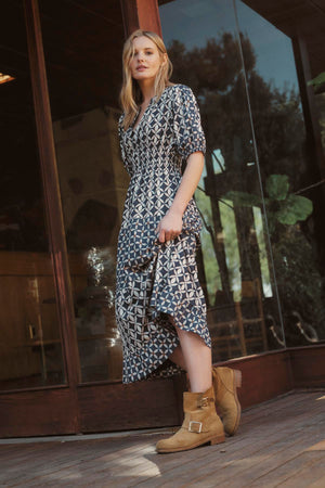 A woman in a JOEY DRESS by Velvet by Graham & Spencer, featuring a patterned, printed cotton cambric fabric and a v-neckline, stands in front of a glass door while holding the edge of her dress. She is wearing brown boots, and there are plants visible through the glass.