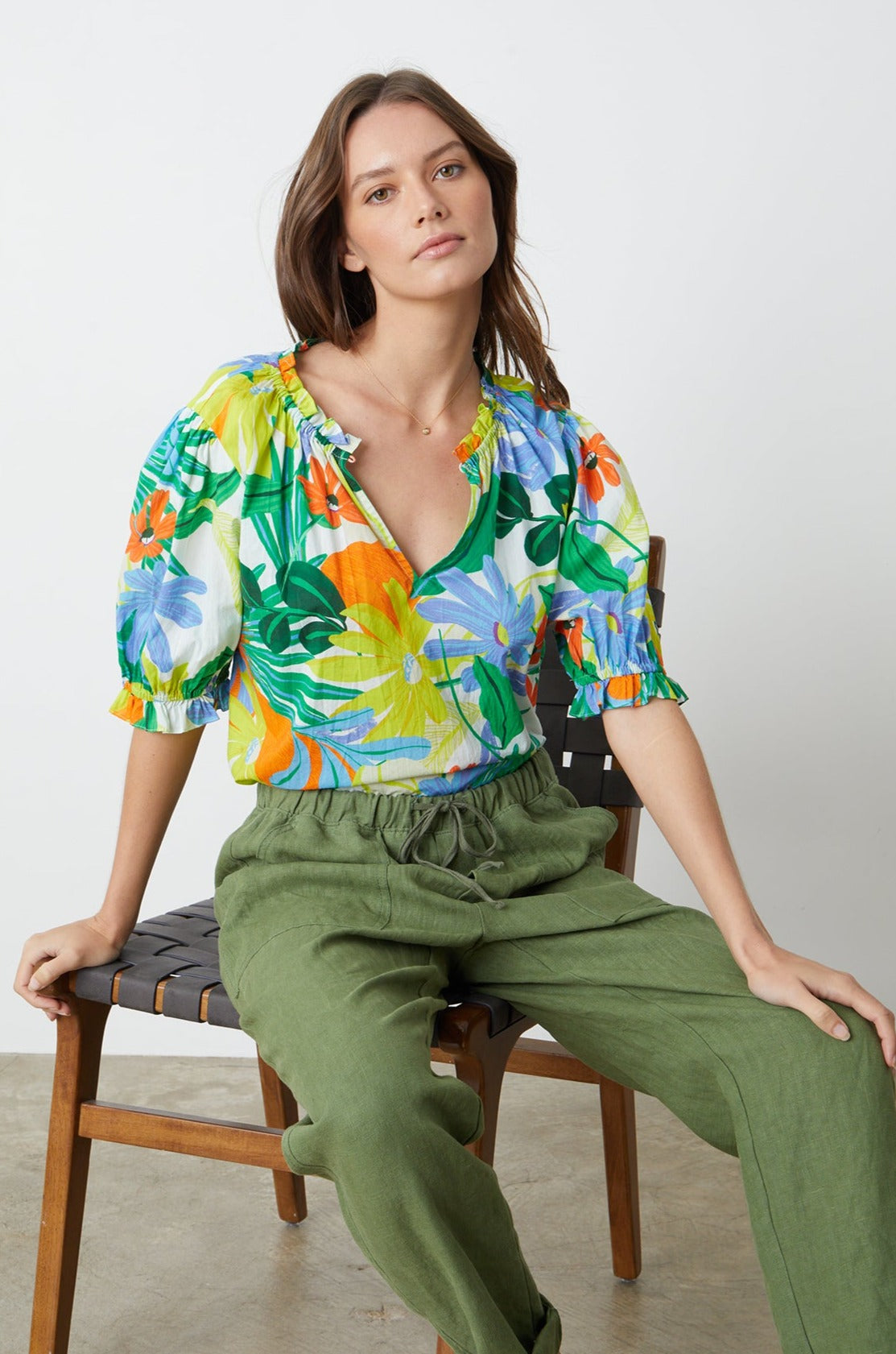   A woman in a CARRIE PRINTED BOHO TOP by Velvet by Graham & Spencer and green pants is sitting on a chair. 