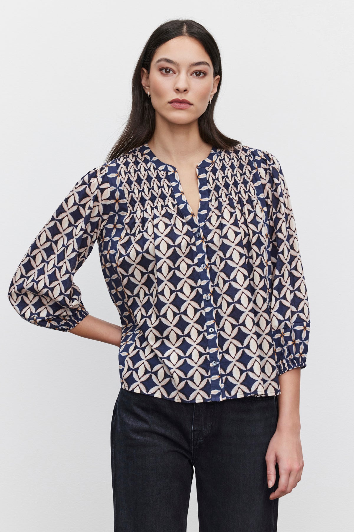   A woman with long dark hair wears a blue and beige geometric-patterned HADDON TOP from Velvet by Graham & Spencer, featuring smocked details, paired with dark jeans. She stands against a plain white background, looking straight ahead with a neutral expression. 