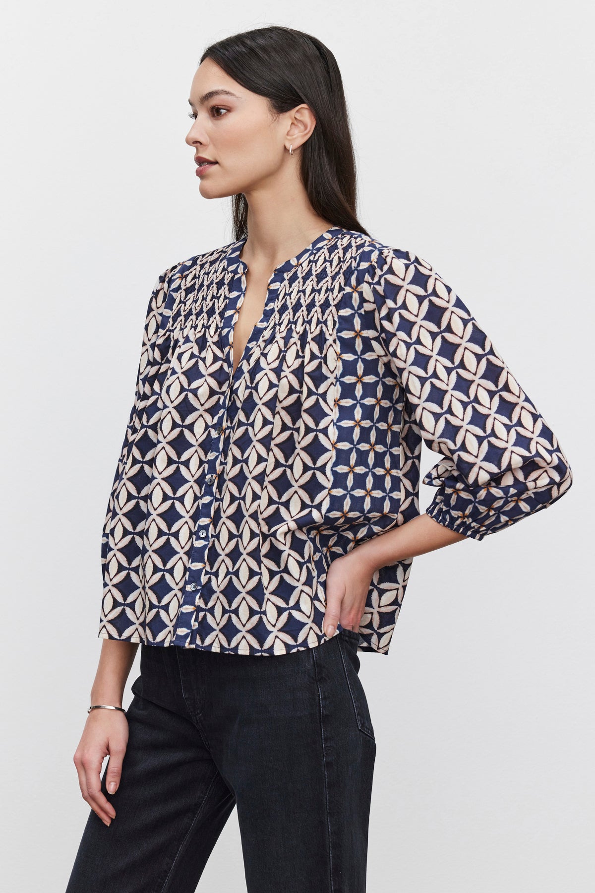   A woman with long dark hair is wearing the HADDON TOP by Velvet by Graham & Spencer, which features a geometric design, and dark pants. She is standing sideways with one hand on her hip against a plain white background. 