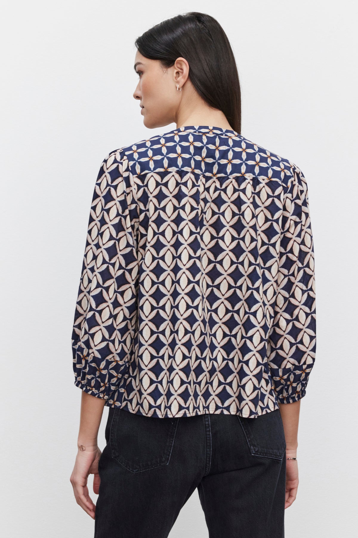   A person is wearing the HADDON TOP by Velvet by Graham & Spencer, featuring three-quarter sleeves and a patterned design, along with dark jeans. They stand with their back facing the camera, highlighting the delicate smocked details of the blouse. 