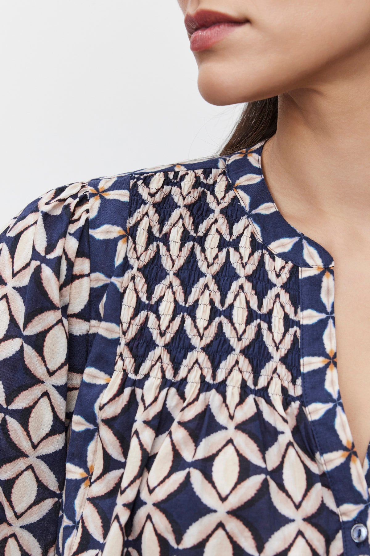   Close-up of a woman wearing the Haddon Top by Velvet by Graham & Spencer, a blue patterned blouse with white geometric designs. Only the lower half of her face and the top part of the blouse, which features delicate smocked details and is made from printed cotton cambric fabric, are visible. 