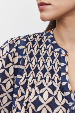 Close-up of a woman wearing the Haddon Top by Velvet by Graham & Spencer, a blue patterned blouse with white geometric designs. Only the lower half of her face and the top part of the blouse, which features delicate smocked details and is made from printed cotton cambric fabric, are visible.