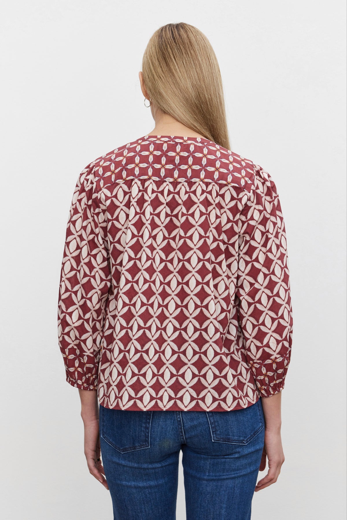   A person with long blonde hair is shown from the back, wearing the HADDON TOP by Velvet by Graham & Spencer—a red and white printed cotton cambric blouse with three-quarter sleeves—and blue jeans, exuding casual refinement. 