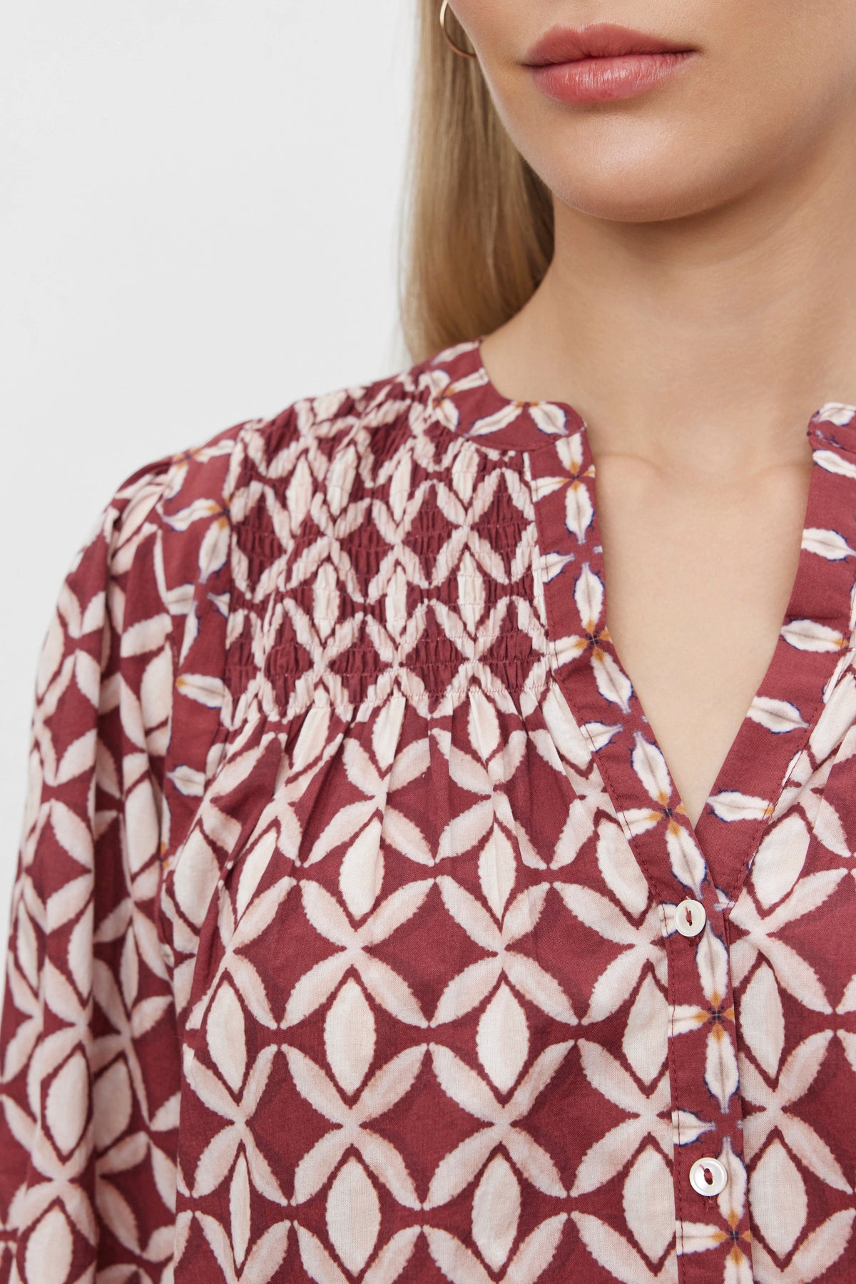   Close-up of a person wearing the HADDON TOP from Velvet by Graham & Spencer, a maroon and white geometric patterned blouse with smocked detail near the neckline and buttoned front. This piece, crafted from printed cotton cambric, exudes casual refinement against a plain white background. 