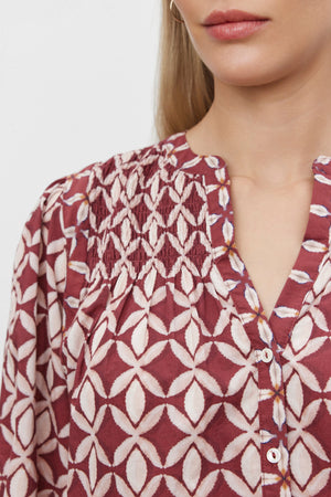 Close-up of a person wearing the HADDON TOP from Velvet by Graham & Spencer, a maroon and white geometric patterned blouse with smocked detail near the neckline and buttoned front. This piece, crafted from printed cotton cambric, exudes casual refinement against a plain white background.