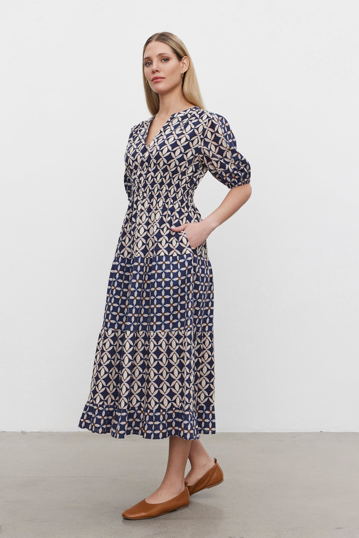   A person stands against a plain background wearing the JOEY DRESS from Velvet by Graham & Spencer, featuring a blue and white print in cotton cambric fabric with a v-neckline. They are paired with brown flats and have their hands in the dress pockets. 