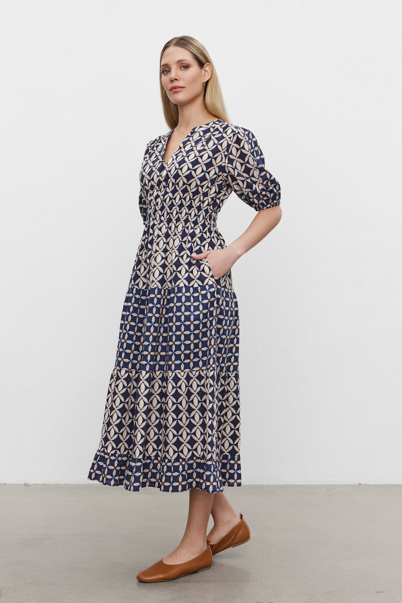 A person stands against a plain background wearing the JOEY DRESS from Velvet by Graham & Spencer, featuring a blue and white print in cotton cambric fabric with a v-neckline. They are paired with brown flats and have their hands in the dress pockets.