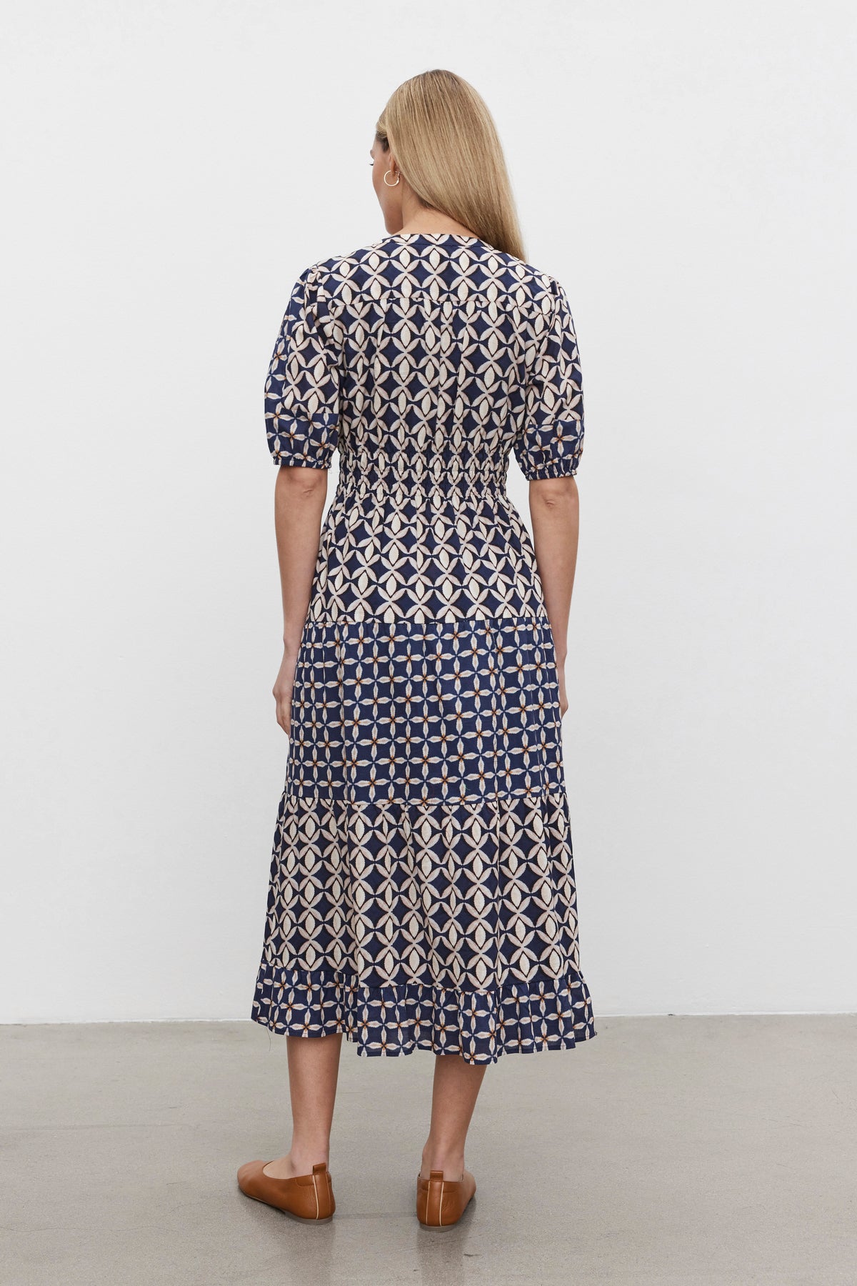   A person with long hair is standing facing away, wearing the JOEY DRESS by Velvet by Graham & Spencer, featuring a patterned geometric design and a two-tiered skirt, paired with brown shoes. 