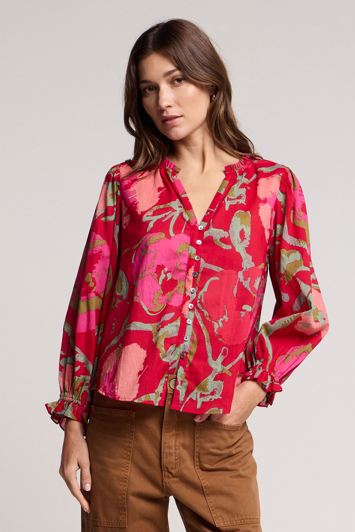 A woman wears the AVENA TOP by Velvet by Graham & Spencer, a long-sleeved red and pink floral button-up blouse, paired with brown pants and standing with one hand in her pocket.