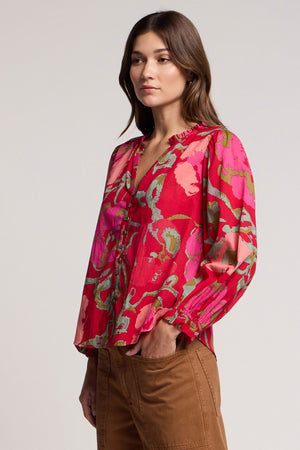 A woman wears the AVENA TOP by Velvet by Graham & Spencer—a vibrant pink and red floral long-sleeve blouse—with brown pants, standing with one hand in her pocket against a plain background.