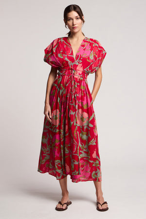 A woman wears the LEILANI DRESS by Velvet by Graham & Spencer, featuring a rose print with red and pink florals, short sleeves, and sandals against a plain background.