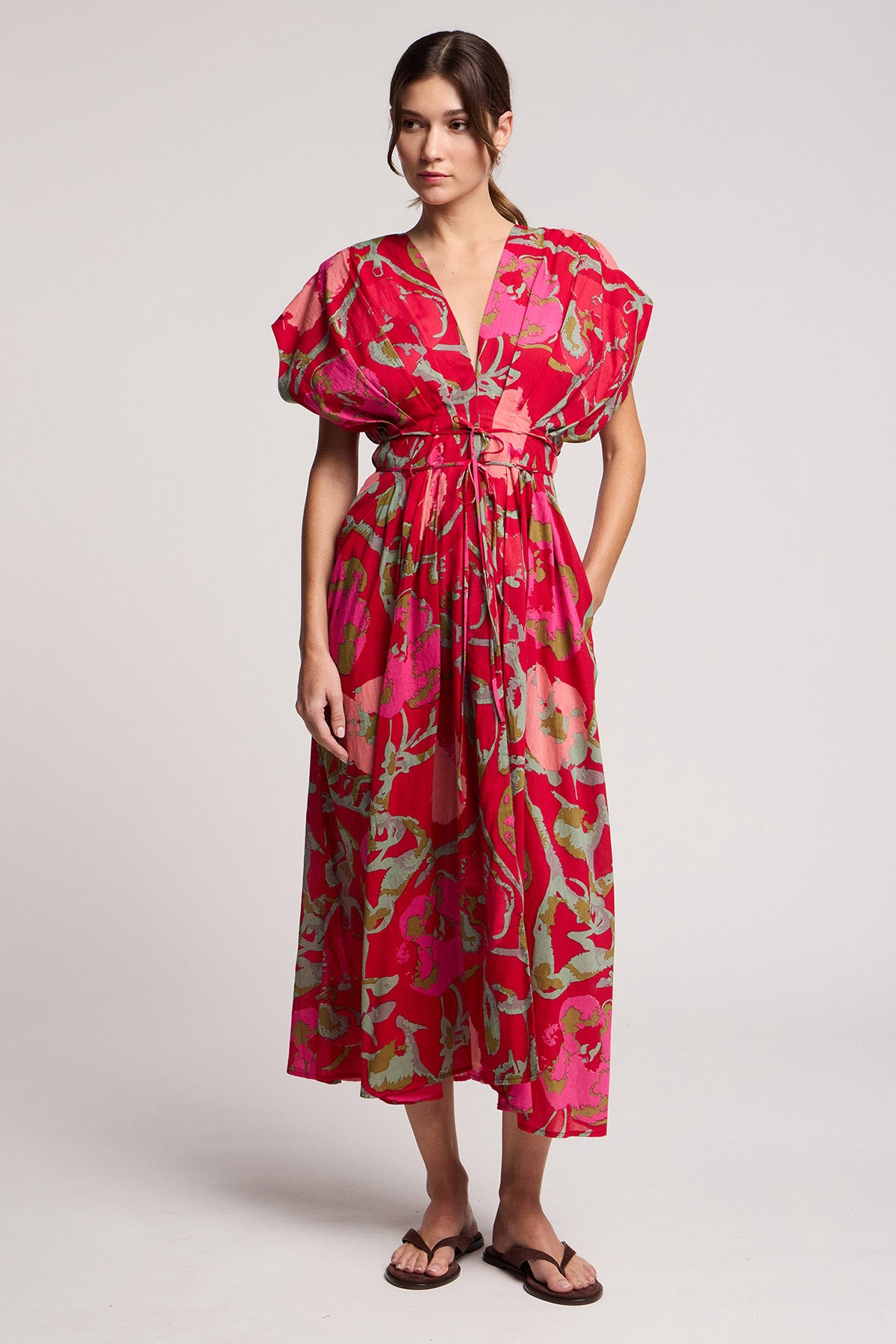 The LEILANI DRESS by Velvet by Graham & Spencer features a rose print with red and pink florals, short sleeves, a belt, and falls to midi length—styled simply with sandals against a plain background.