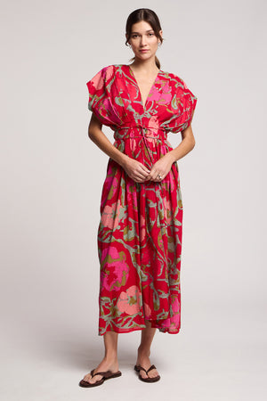 A woman stands against a plain background in the LEILANI DRESS by Velvet by Graham & Spencer—a loose-fitting, rose print dress with short sleeves—paired with sandals.