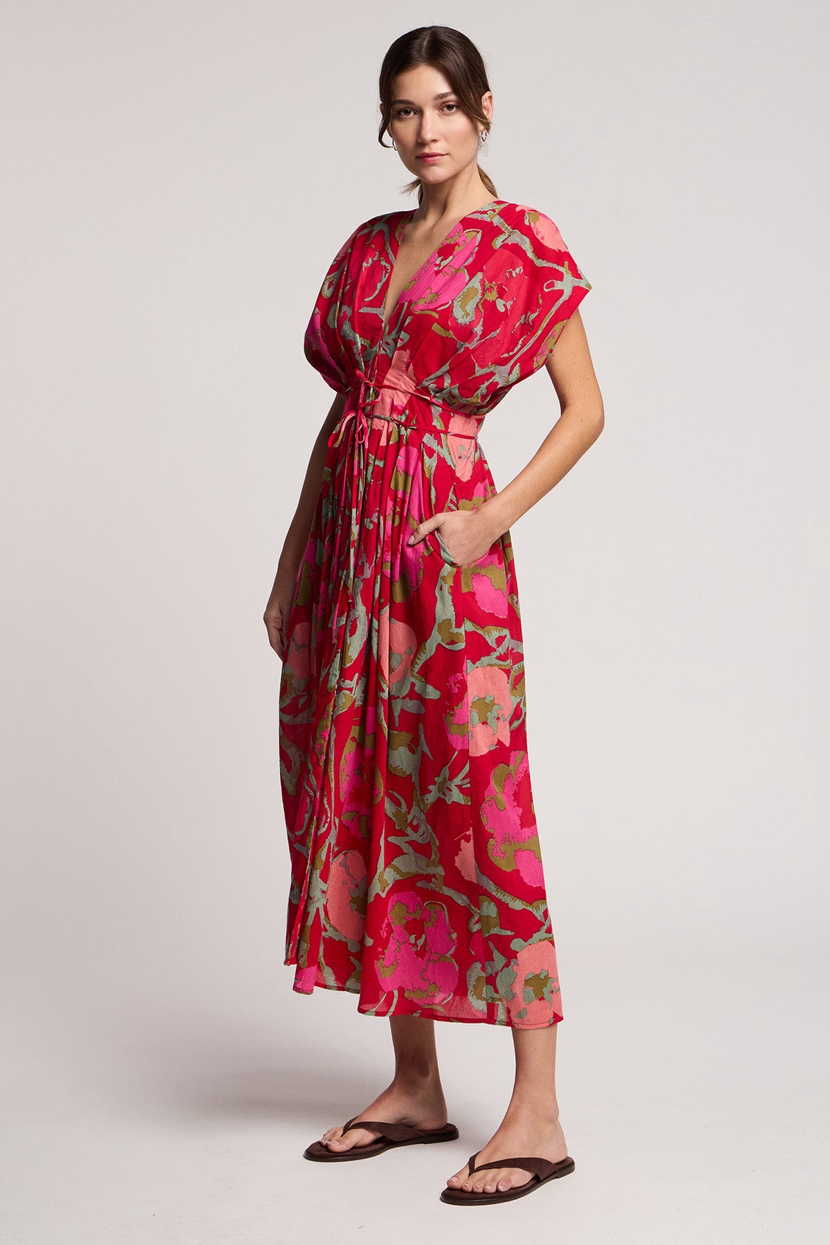A woman wears the LEILANI DRESS by Velvet by Graham & Spencer—a pink rose print midi dress with short sleeves and a V-neck—paired with black sandals, standing against a plain light background.