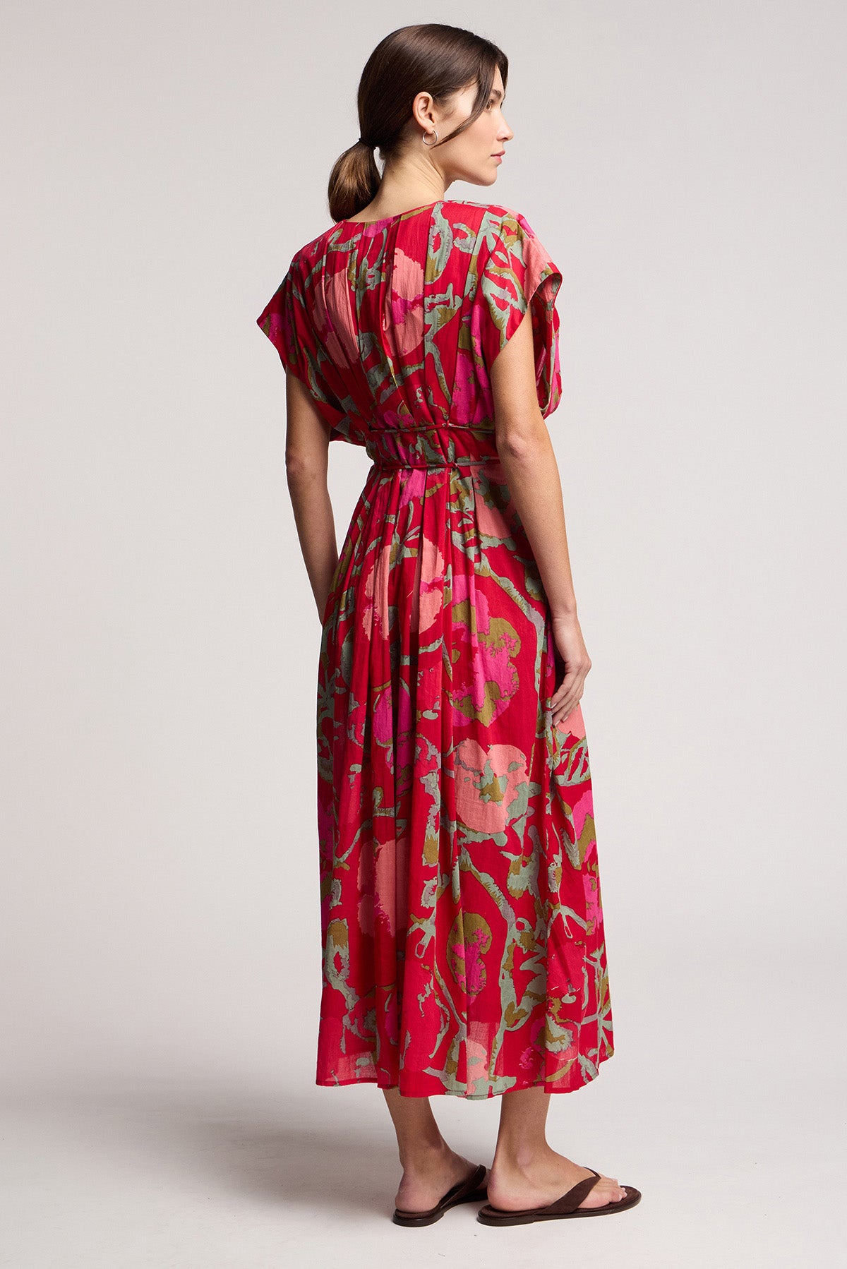 A woman faces away, wearing the LEILANI DRESS by Velvet by Graham & Spencer—a red and green rose print dress with a cinched waist and short sleeves—paired with brown sandals against a plain background.