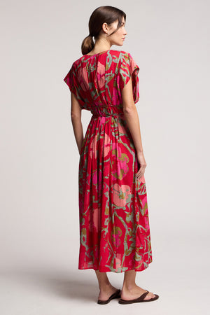 A woman faces away, wearing the LEILANI DRESS by Velvet by Graham & Spencer—a red and green rose print dress with a cinched waist and short sleeves—paired with brown sandals against a plain background.