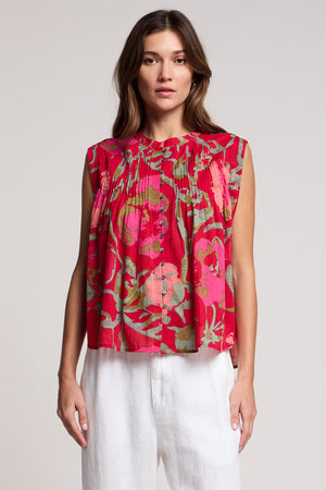 A woman wears the JEANNE TOP by Velvet by Graham & Spencer—a romantic sleeveless rose vine blouse with pleats—paired with white pants, standing against a plain light background.