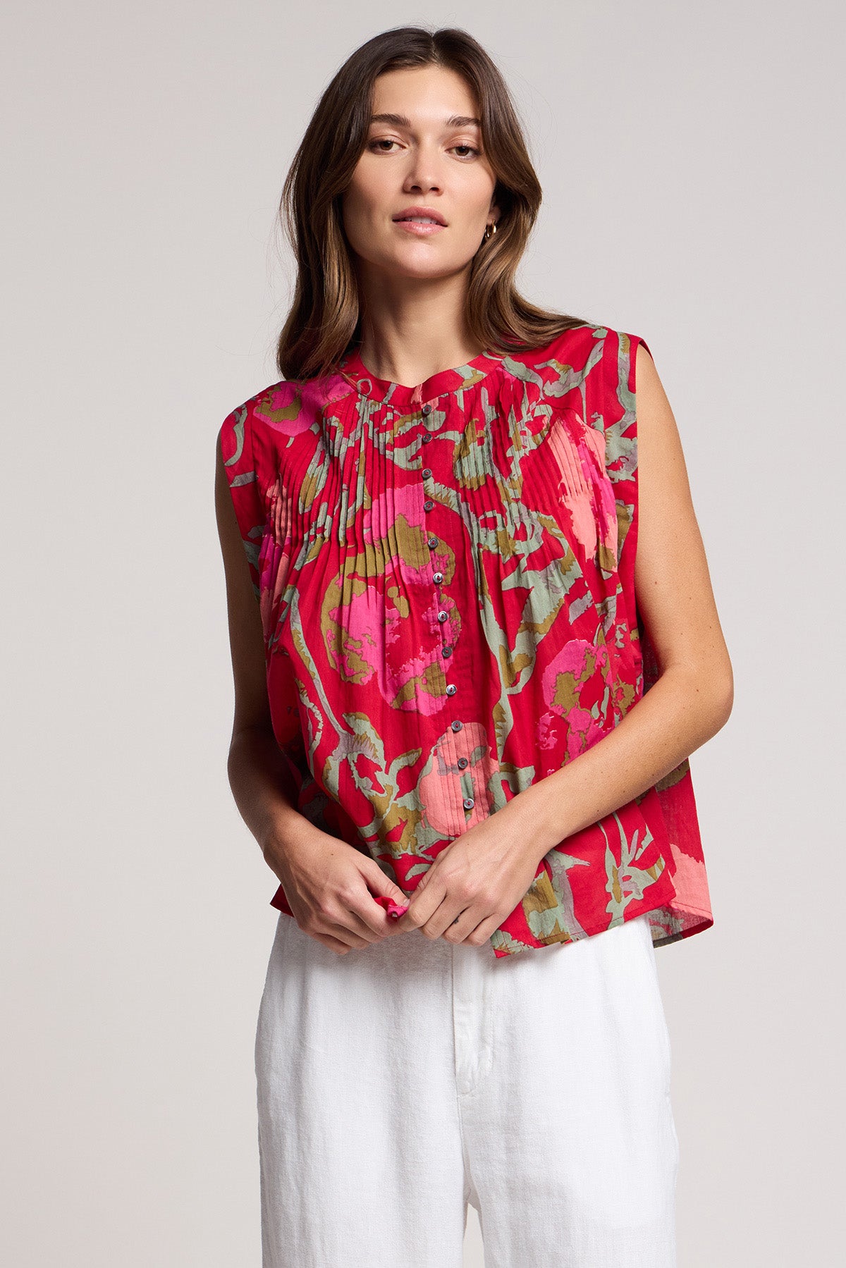 A woman wearing the Velvet by Graham & Spencer JEANNE TOP, a romantic sleeveless blouse with red floral pleats, and white pants stands against a plain light background.