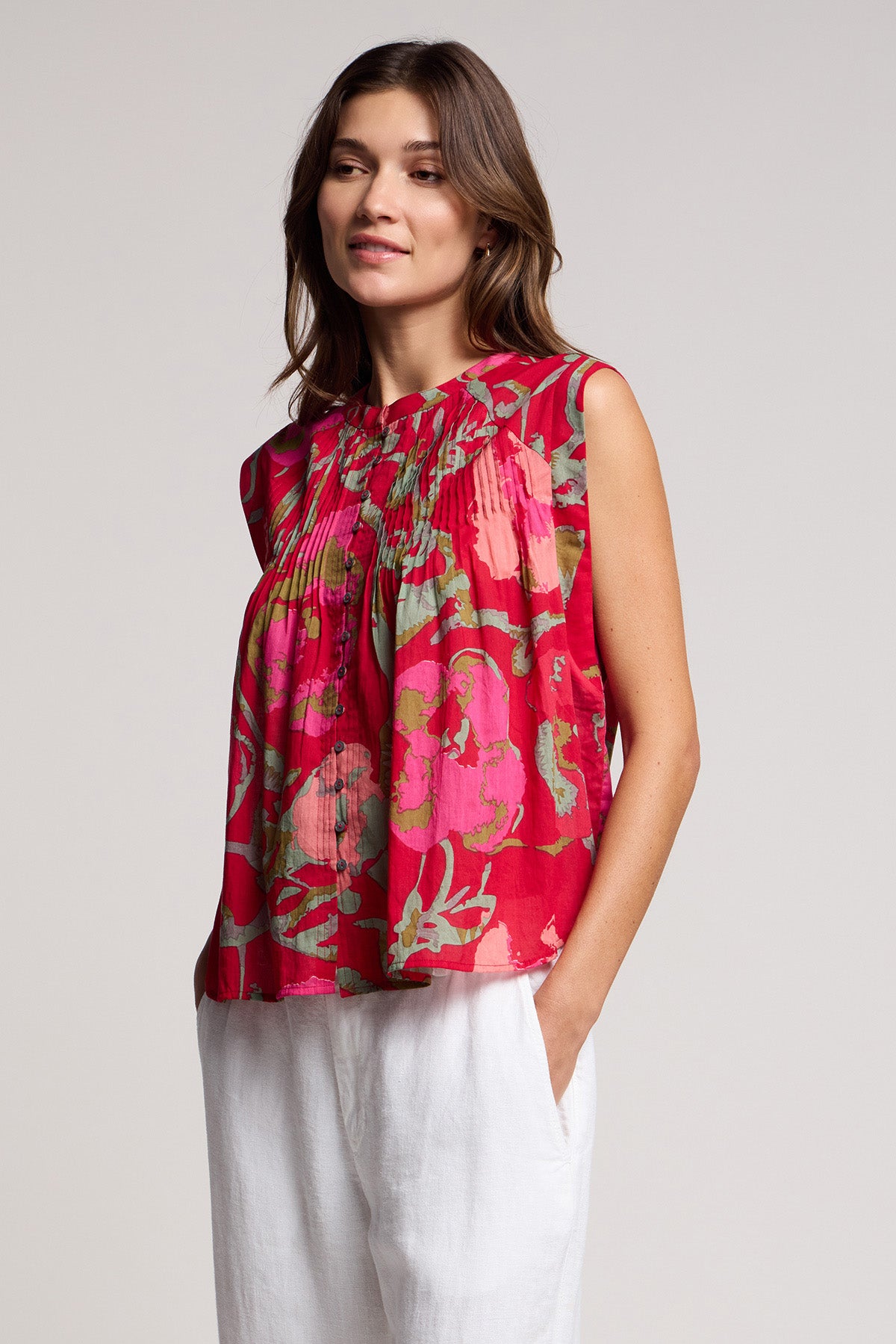 A woman wears the JEANNE TOP by Velvet by Graham & Spencer, a romantic sleeveless blouse in a red rose vine print, paired with white pants and standing with one hand in her pocket against a plain background.-41527348986049