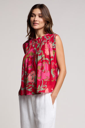 A woman wears the JEANNE TOP by Velvet by Graham & Spencer, a romantic sleeveless blouse in a red rose vine print, paired with white pants and standing with one hand in her pocket against a plain background.