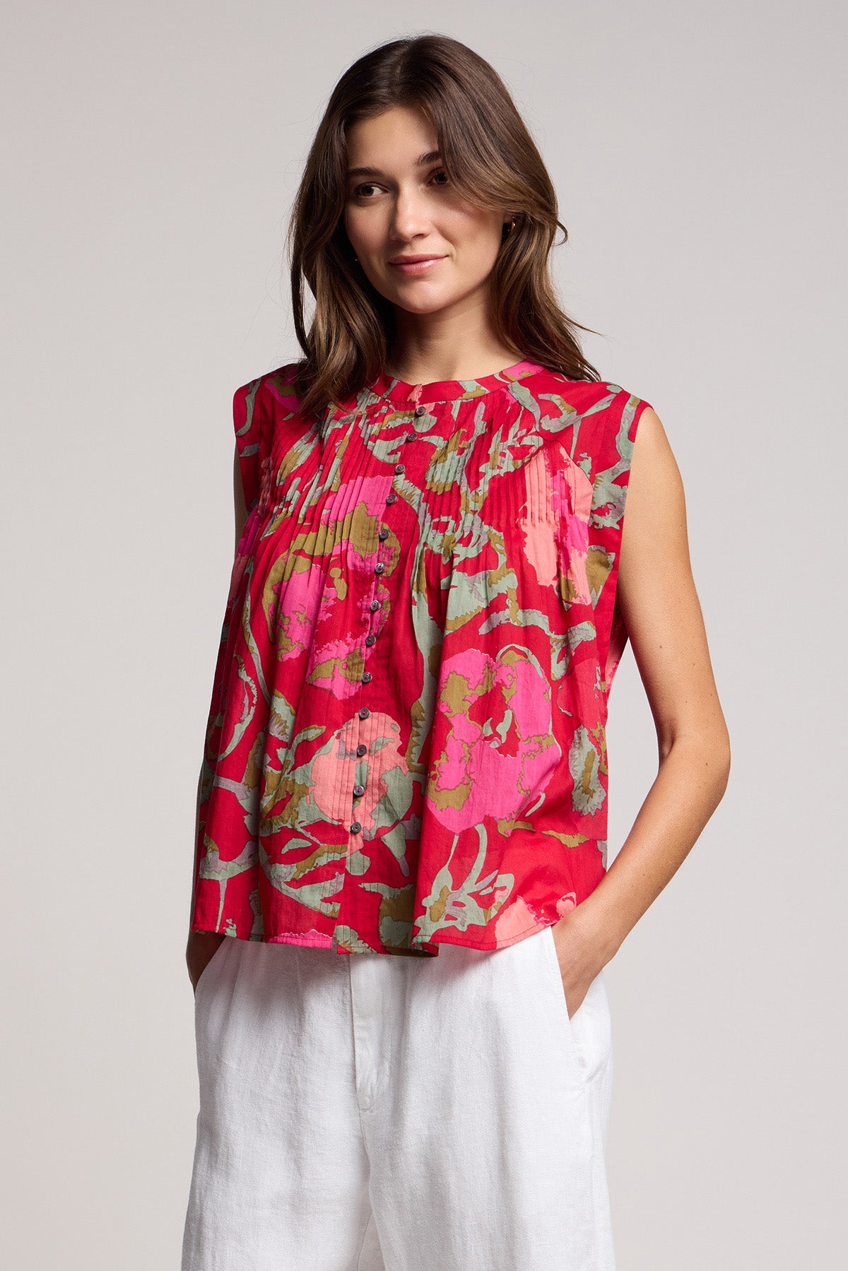 Wearing the JEANNE TOP by Velvet by Graham & Spencer—a sleeveless red floral blouse—paired with white pants, a person stands with hands in pockets against a plain background.