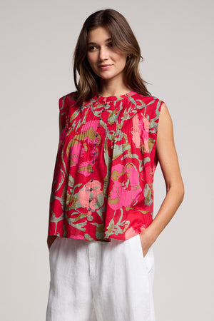 Wearing the JEANNE TOP by Velvet by Graham & Spencer—a sleeveless red floral blouse—paired with white pants, a person stands with hands in pockets against a plain background.