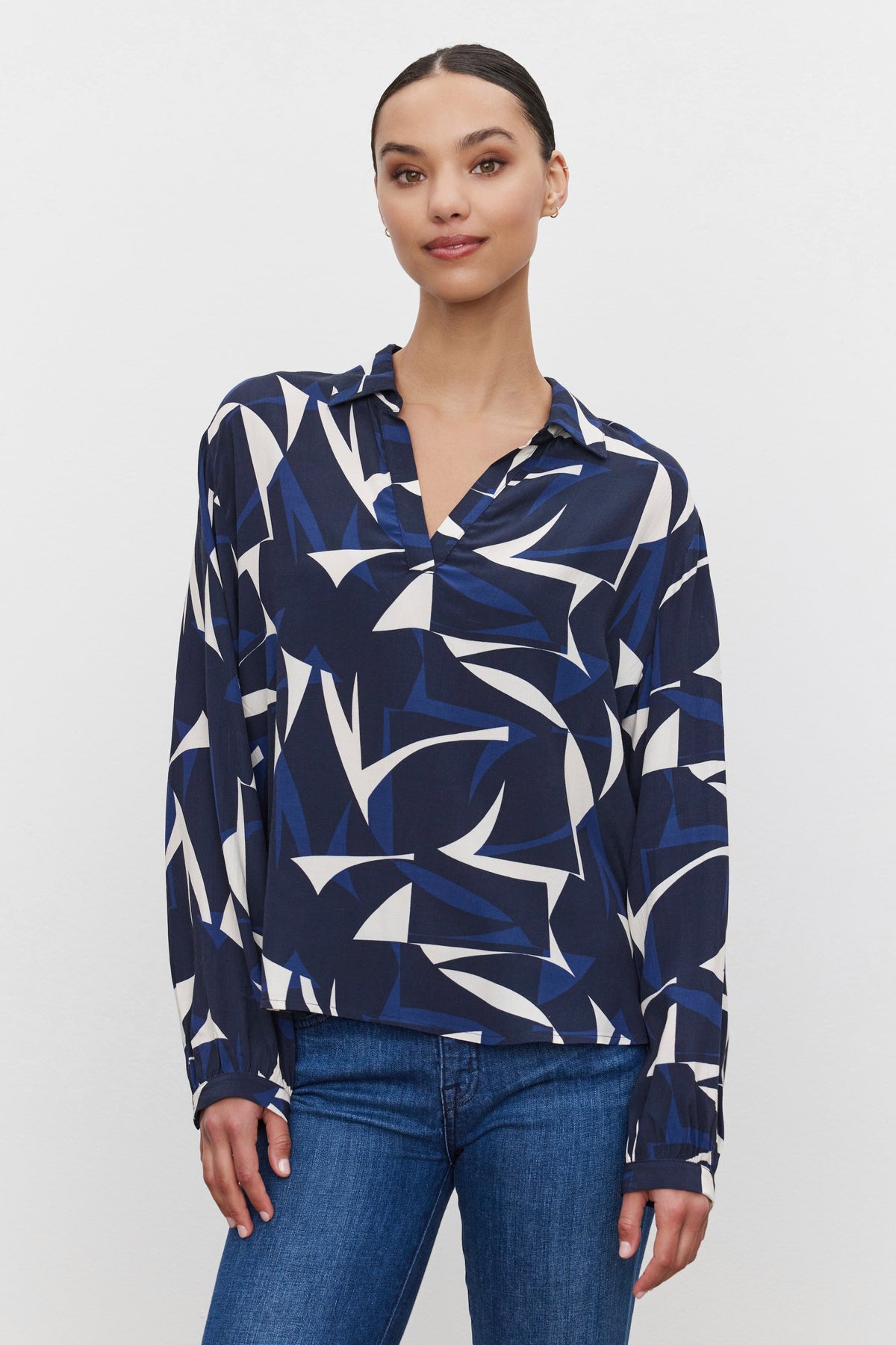 A person stands against a plain background, wearing the Velvet by Graham & Spencer CARAH TOP, a long-sleeve, split-neck blouse in blue, white, and black geometric shapes made from printed royal crepe fabric, paired with blue jeans.-37676902056129