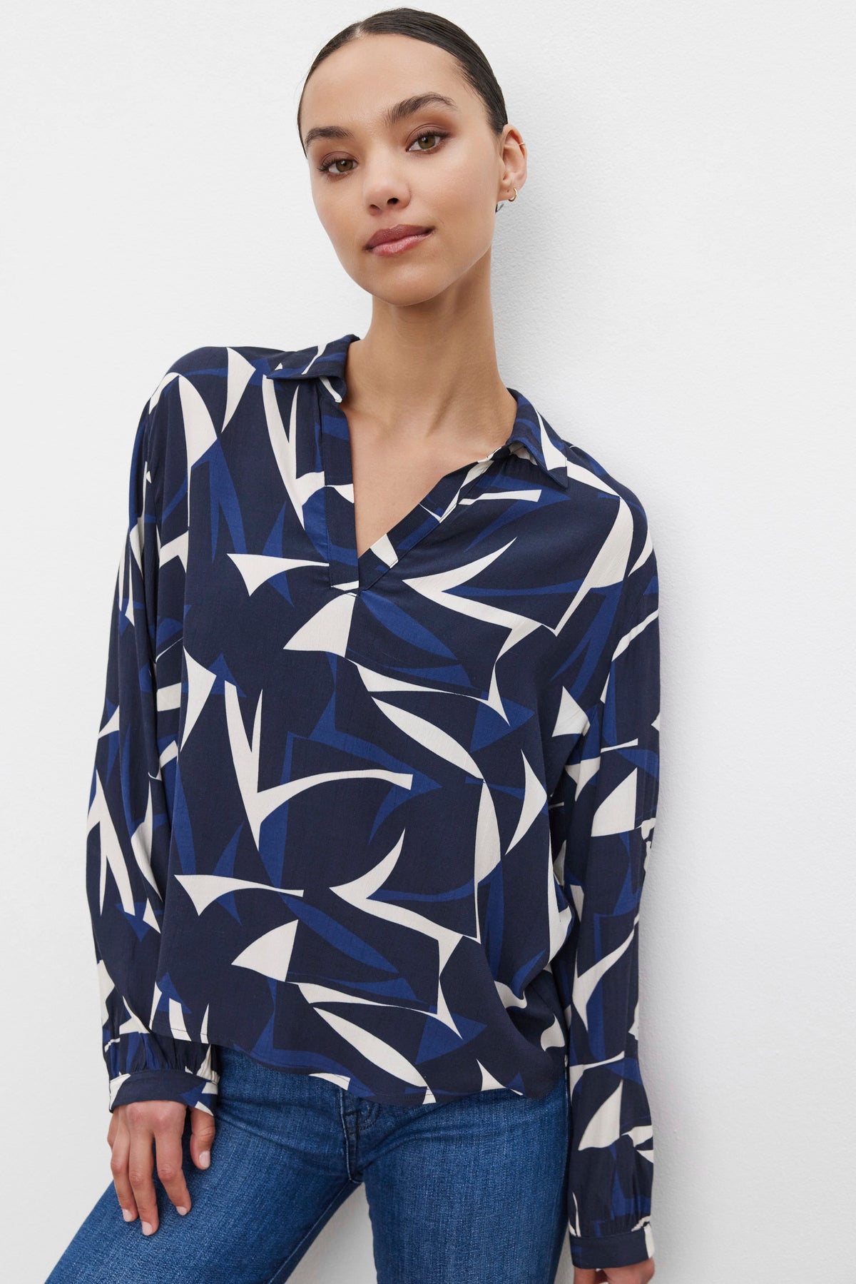   Person wearing the CARAH TOP from Velvet by Graham & Spencer, which features a blue and white patterned blouse with a split neck and textured fabric, paired with jeans, stands against a plain white background. 